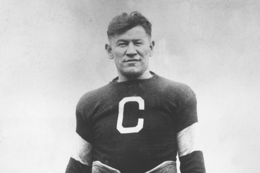 A portrait of American athlete Jim Thorpe posing in a football uniform on a field.