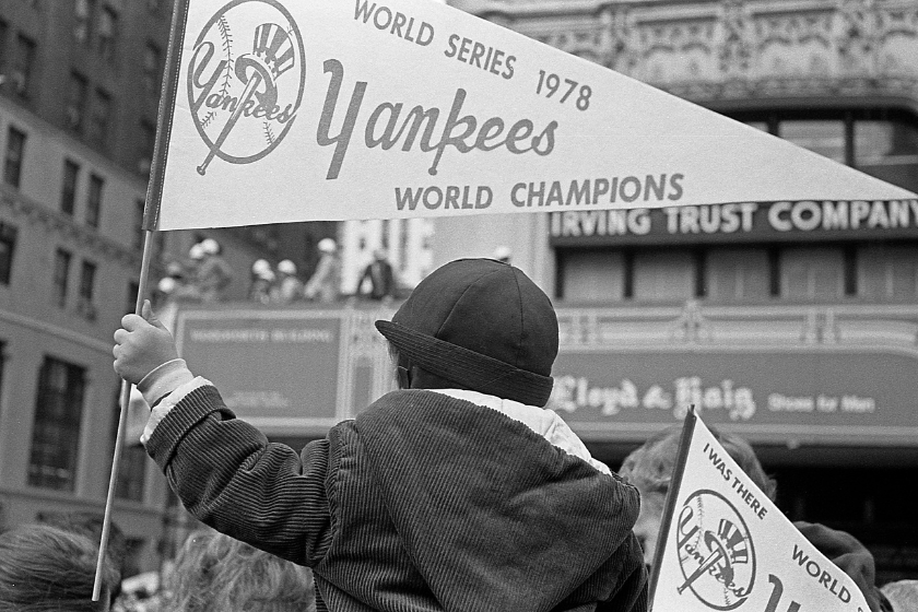 A young boy sits on his fathers shoulders and holds a Yankees banner at a ticker tape parade