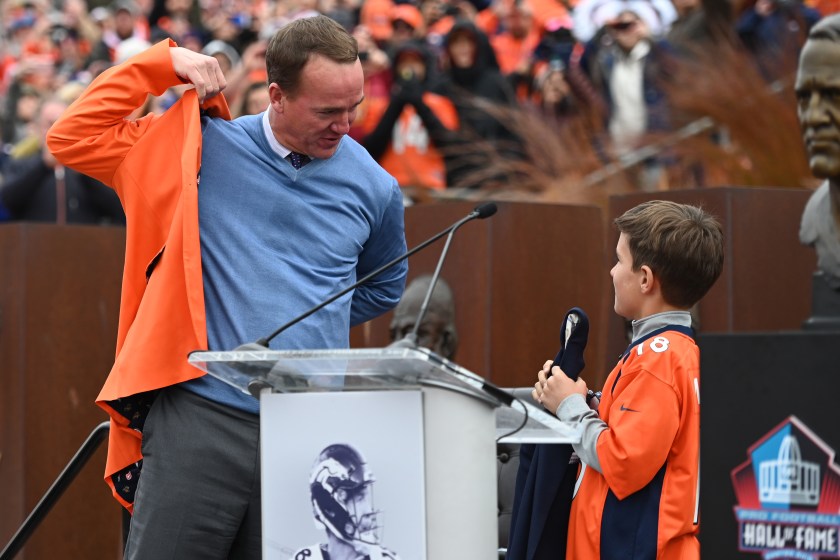 Marshall Manning hands Peyton Manning an orange jacket.